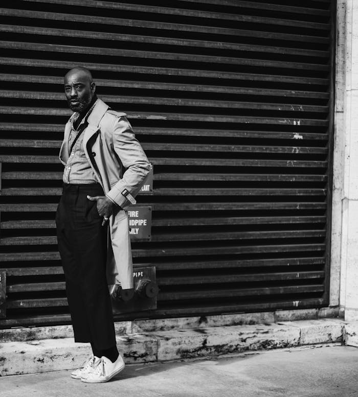 Black and white full body of African American bold male looking at camera while standing  on tiptoes on street near wall with horizontal boards with hands in pockets